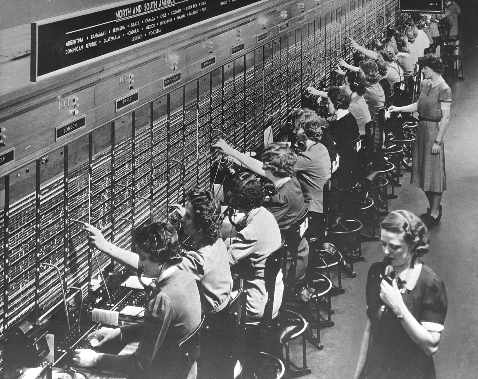 Schwarzweißfoto: eine Reihe Telefonistinnen stöpselt Gespräche an einer langen Vermittlungswand